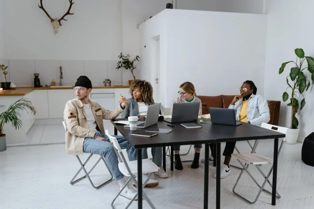 En gruppe unge professionelle arbejder sammen omkring et sort bord i et lyst kontormiljø med bærbare computere, planter og et åbent køkken i baggrunden.